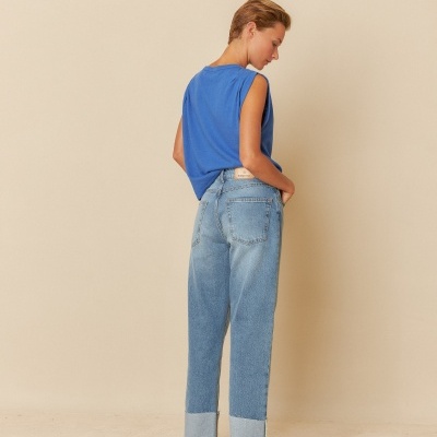 Modelo de costas com calças de ganga azul claro e t-shirt azul sem mangas, fundo bege.