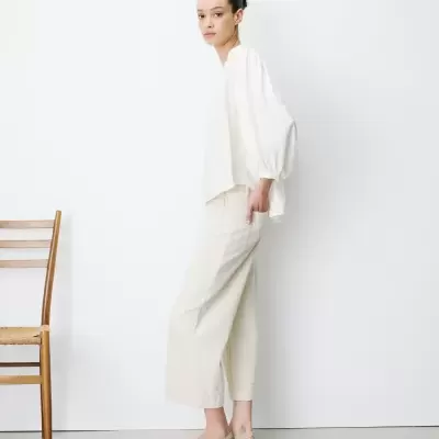 Mulher de perfil com blusa branca de mangas bufantes e calças creme largas, sapatos rasos beges, cadeira de madeira ao lado.