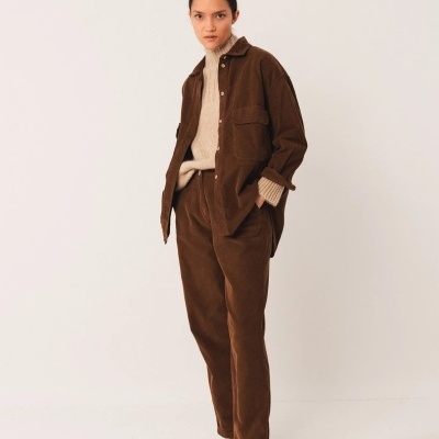 Mulher com casaco e calças de veludo castanho escuro, camisola bege e sapatos castanhos em fundo branco.