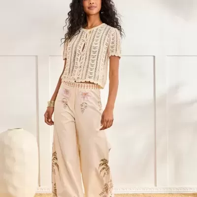 Mulher com camisa de malha bege e calças largas creme bordadas, em ambiente interior neutro