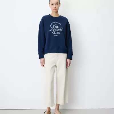 Pessoa com sweatshirt azul escuro escrito THE Lovers CLUB, calças creme e sapatos bege