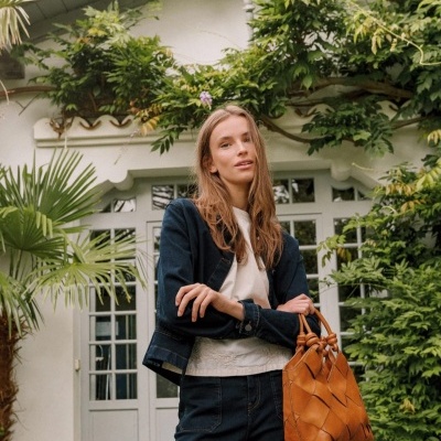 Mulher com casaco azul escuro e mala castanha em padrão trançado, em frente a porta branca e plantas verdes