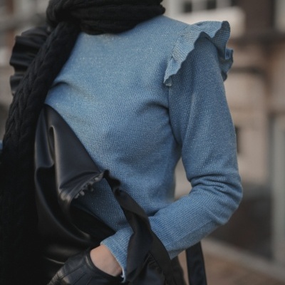 Mulher com blusa azul clara de malha com folhos, cachecol e luvas pretas, casaco preto, fundo urbano desfocado