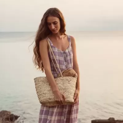 Mulher com vestido lilás e branco segurando mala de palma à beira-mar ao entardecer