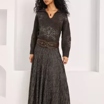 Mulher com camisola castanha bordada, saia plissada cinzenta, cinto largo castanho e botas castanhas.