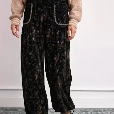Pantalonas pretas largas com padrão floral subtil e bordados nos bolsos, sweater bege e ténis bege em fundo branco.
