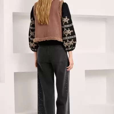 Mulher de costas com colete castanho de pelo, camisa preta bordada, calças pretas largas com strass e sapatos castanhos