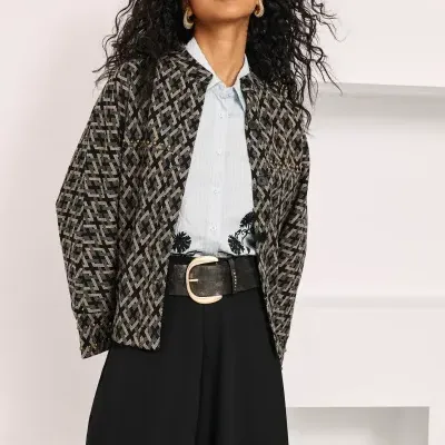 Mulher vestindo casaco preto e cinza estampado, camisa azul clara, saia e cinto preto com fivela dourada.