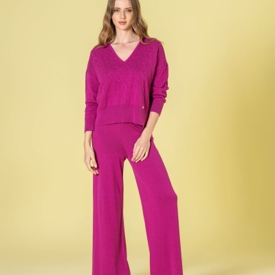 Mulher a usar conjunto de malha magenta com blusa de manga comprida e calças largas, sandálias castanhas, fundo amarelo.