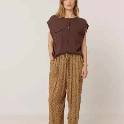Mulher de colete castanho e calças largas castanhas com padrão em fundo neutro