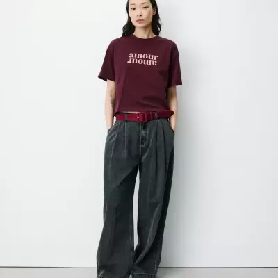Mulher vestindo t-shirt vinho com texto amour amour, calças cinza largas, cinto e sapatos vinho, em fundo branco.