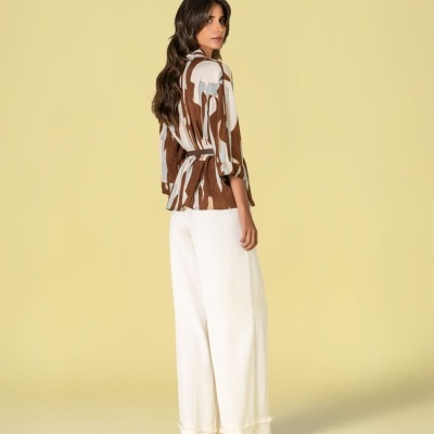 Mulher com blusa padrão castanho e branco e calças largas brancas, em fundo amarelo.
