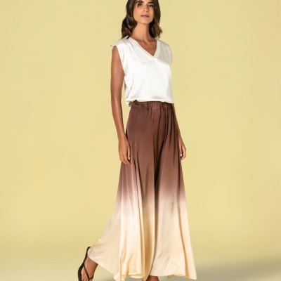 Mulher com blusa branca sem mangas e calças largas castanho e bege degradê com sandálias pretas.