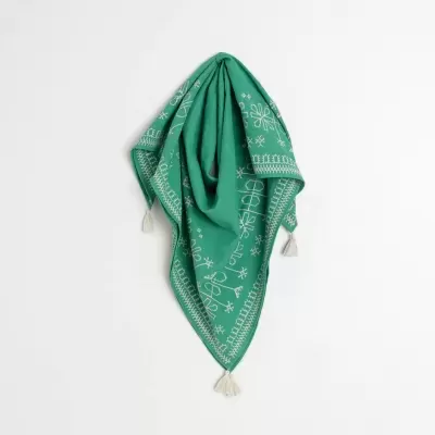 Lenço triangular verde com bordados e tasséis brancos pendurado numa parede branca