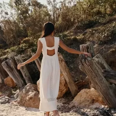 Mulher de costas usando vestido longo branco com costas abertas em ambiente exterior arenoso com pedras e troncos