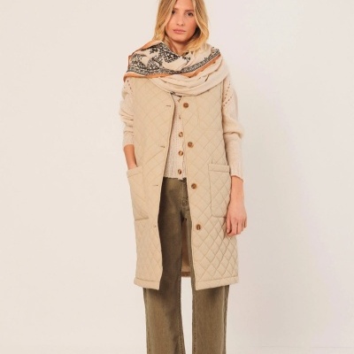 Mulher com colete bege acolchoado, camisola e calças castanhas, lenço estampado, fundo branco