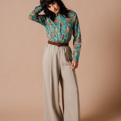 Mulher de pé com camisa floral verde e calças largas bege num fundo branco neutro