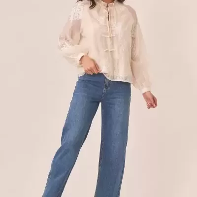 Mulher com blusa transparente bege bordada, calças de ganga azuis e botas castanhas.