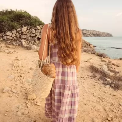 Mulher de vestido aos quadrados lilás e branco a caminhar numa praia com cesta de palha.