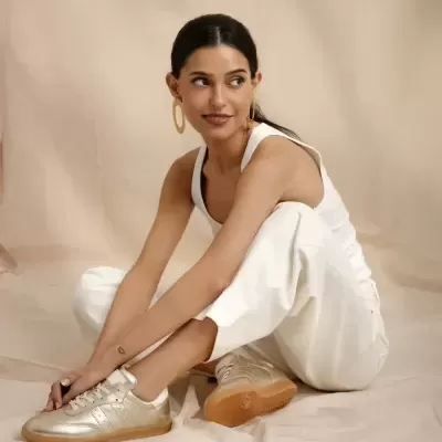 Mulher sentada com calças brancas e ténis dourados numa sala com fundo bege.