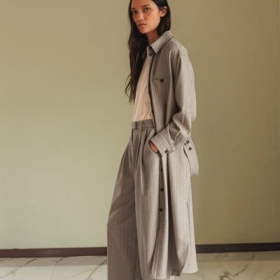 Mulher com conjunto cinza às riscas com casaco comprido e calças largas em fundo bege e chão branco com detalhes cinza.