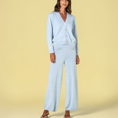 Mulher a usar conjunto azul claro com cardigan e calças largas, e sandálias pretas de tacão, fundo amarelo claro.