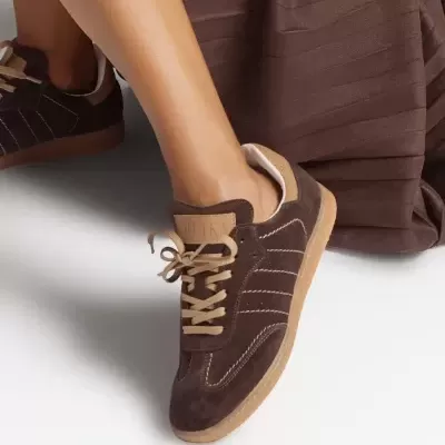 Ténis castanho escuro de camurça com atacadores castanhos claros e etiqueta RUÏKA na língua, junto a saia castanha escura.