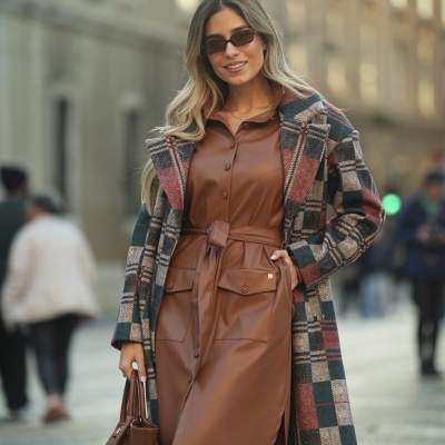Mulher com casaco de padrão xadrez e vestido castanho com cinto, óculos escuros, mala castanha e botas castanhas numa rua.