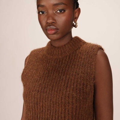 Mulher de pele escura com colete castanho de malha grossa, sem mangas, gola alta canelada