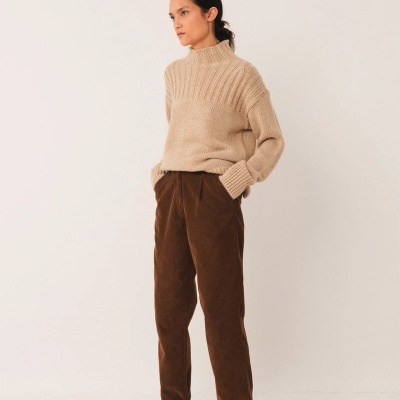 Mulher com camisola bege e calças castanhas em fundo branco