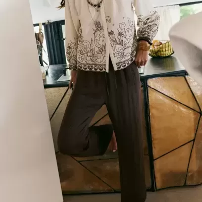 Mulher com blusa bege com padrões florais castanhos e calças castanhas em interior