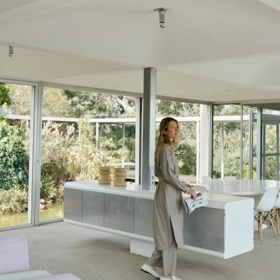 Mulher de roupa cinza clara e ténis brancos num interior moderno com janelas largas e mobiliário branco.