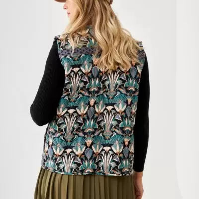 Colete com padrão floral colorido visto de costas, usado com camisola preta, saia verde-oliva plissada e boné bege