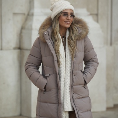 Mulher com casaco acolchoado bege, camisola creme, calças castanhas, gorro com pompom e óculos de sol.