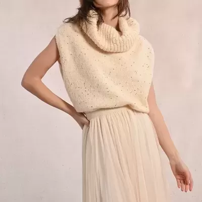 Mulher com top de malha creme e saia comprida plissada creme em fundo claro