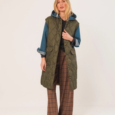 Mulher vestindo colete verde acolchoado comprido, camisa azul com capuz e calças castanhas xadrez num fundo neutro