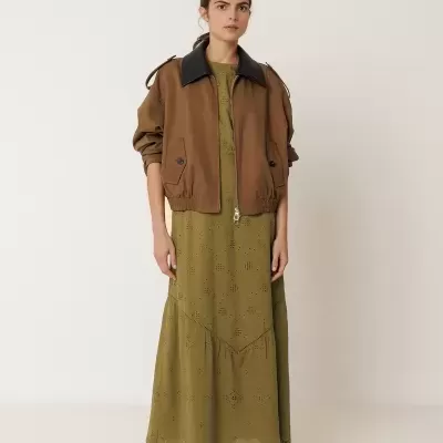 Mulher com vestido verde caqui longo com detalhes vazados e casaco castanho com gola preta.