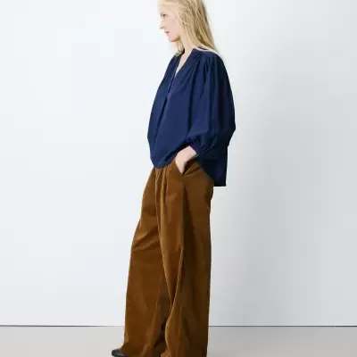 Mulher loira de perfil com blusa azul escura e calças largas de veludo cor de caramelo.