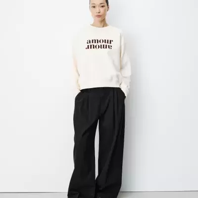 Mulher com sweatshirt branca com texto amour amour e calças pretas largas em fundo branco.