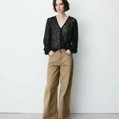 Mulher vestindo blusa preta transparente de renda e calças largas bege em fundo branco.
