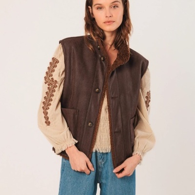 Mulher a usar colete castanho de imitação de pele com interior de pelo, camisola bege bordada e calças de ganga azuis