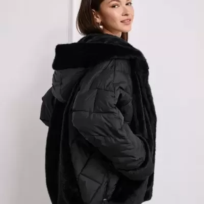 Casaco preto acolchoado com capuz e detalhes em pelúcia, mulher de lado com calças cinza.