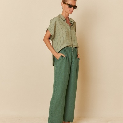 Modelo a usar camisa verde clara de padrão quadriculado e calças largas verdes escuras, com sandálias castanhas e óculos escuros, em fundo bege.