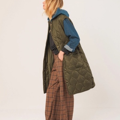 Mulher com colete matelassê verde-oliva, camisa azul, calças xadrez castanhas e sapatos desportivos.