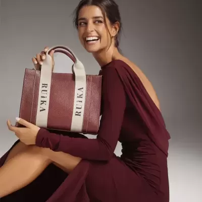 Mulher sentada com vestido bordô e bolsa marrom com alças bege 'RUIKA'.