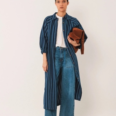 Mulher com casaco azul riscado, t-shirt branca, calças de ganga largas e ténis castanhos, em fundo branco.