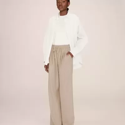 Mulher com camisa branca, camisola branca e calças largas bege com riscas finas.