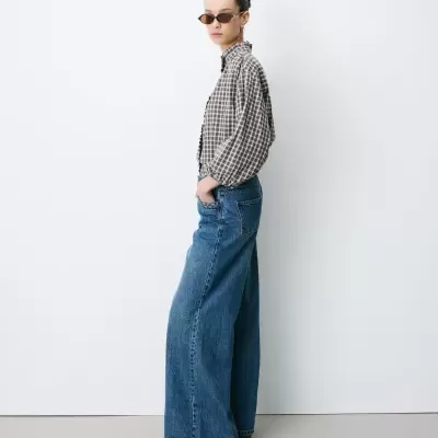 Mulher de perfil com camisa xadrez, calças largas em jeans e óculos de sol