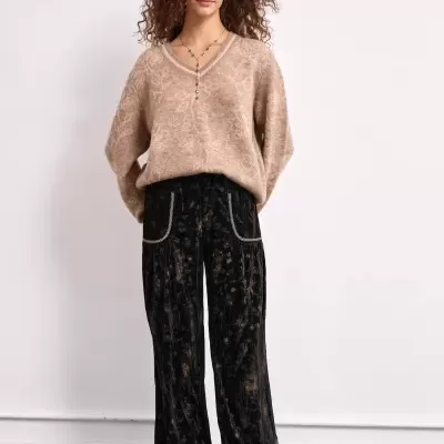 Mulher com camisola bege de malha, calças pretas largas com padrão floral e ténis bege.