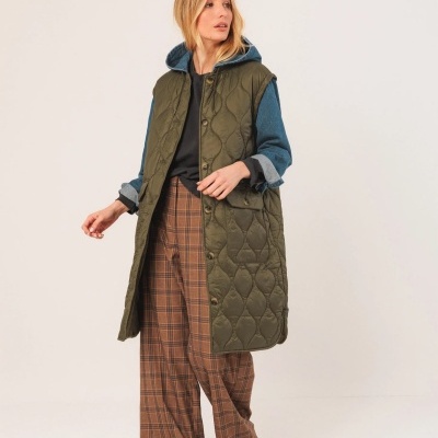 Mulher a usar colete verde-oliva acolchoado comprido com camisola azul, calças castanhas às riscas e sapatos desportivos